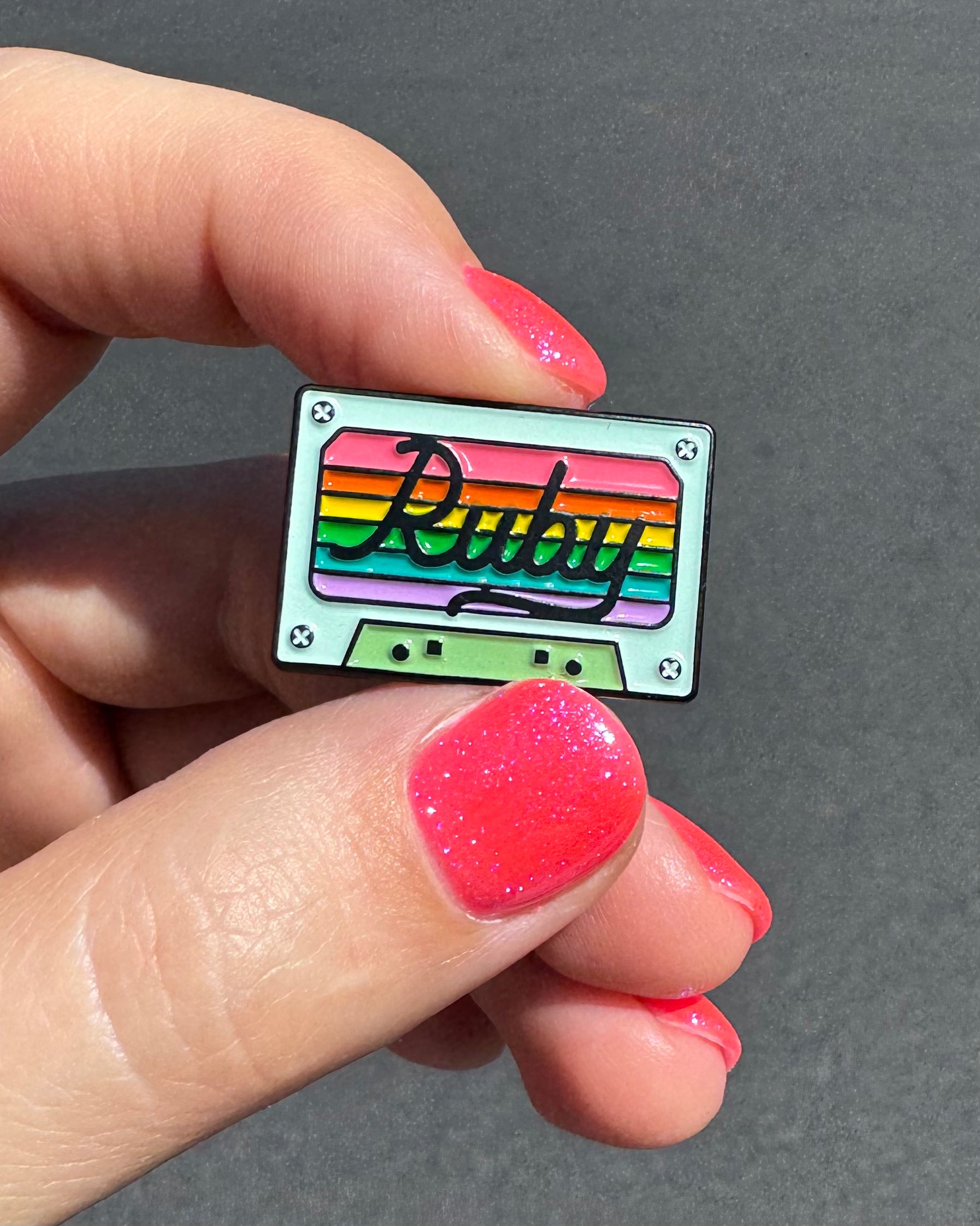 aqua cassette tape pin with rainbow colors, held by a hand with hot pink sparkly nail polish